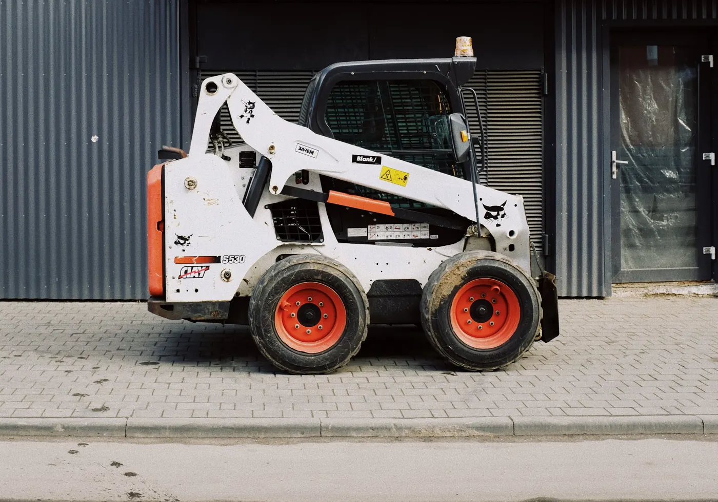 Side view of a white Bobcat skid steer loader with orange wheels, parked in front of a grey industrial building
